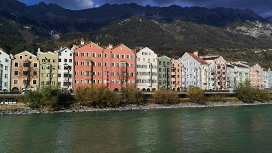 Bunte Häuser am Inn Eine bunte Häuserfront hinter einem Fluss und vor Bergen an einem bewölktem Tag.
